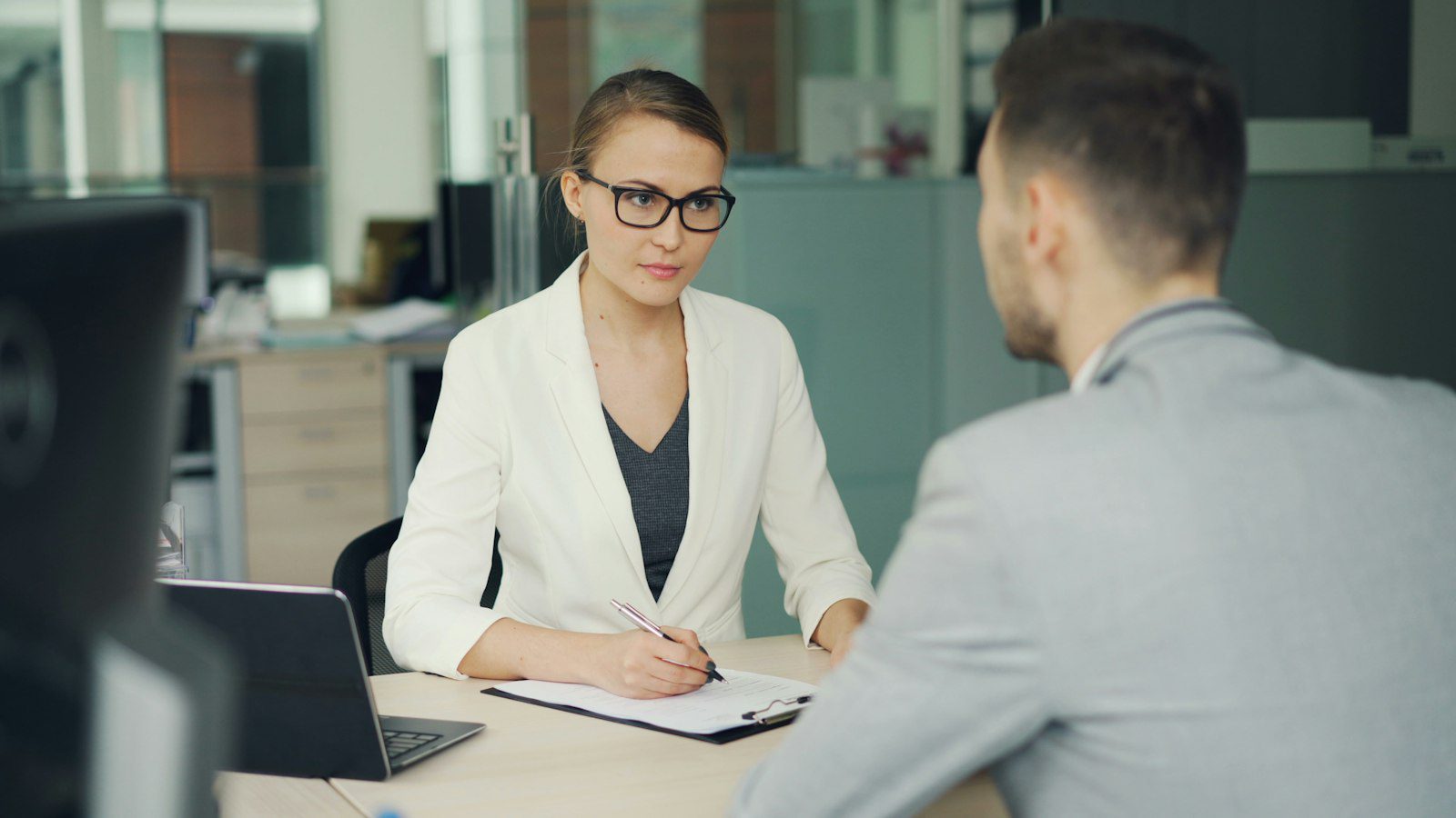 Interviewer in glasses taking notes while a job candidate answers questions across an office desk