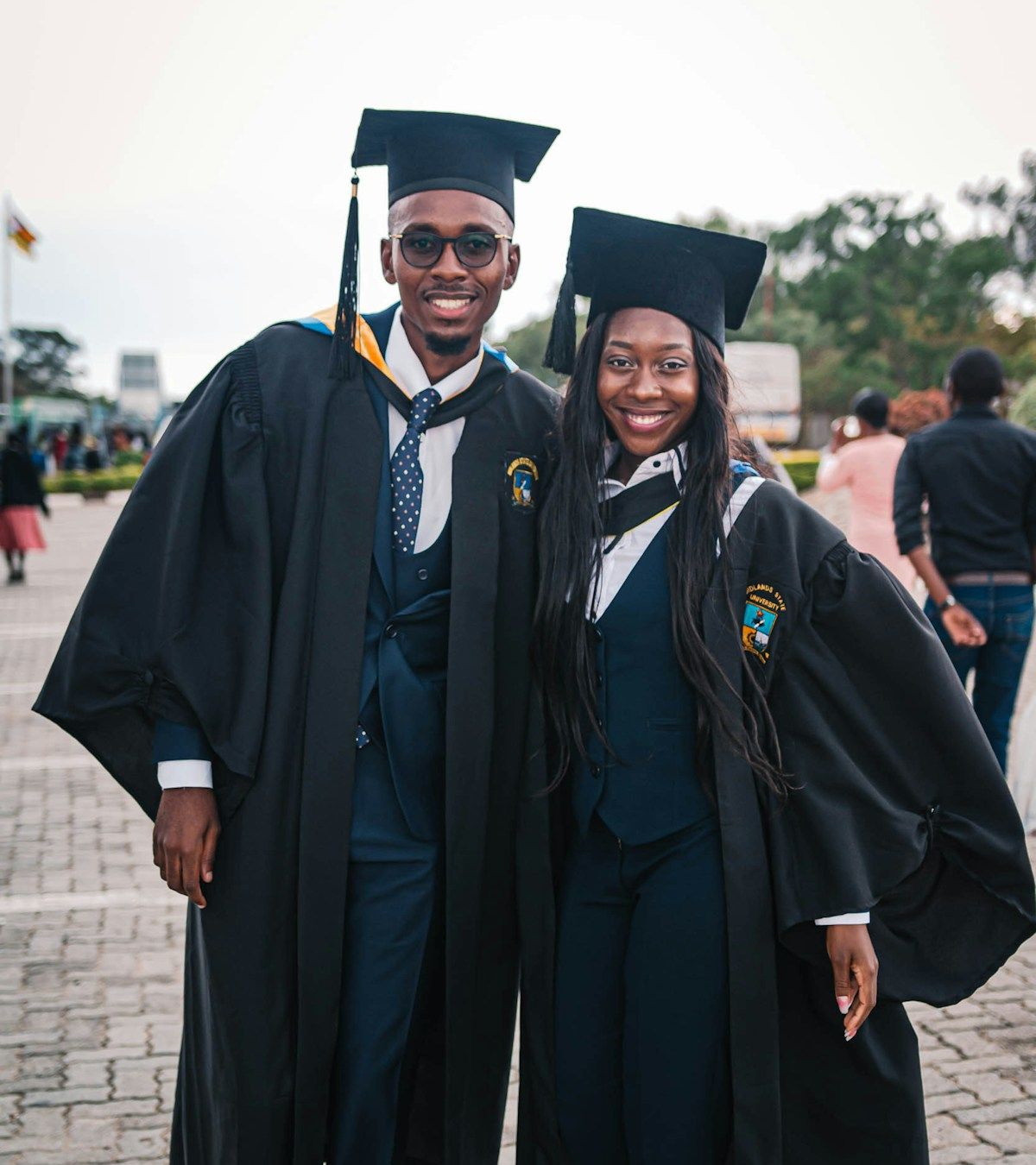 Two smiling young graduates in black academic gowns and caps standing together on campus